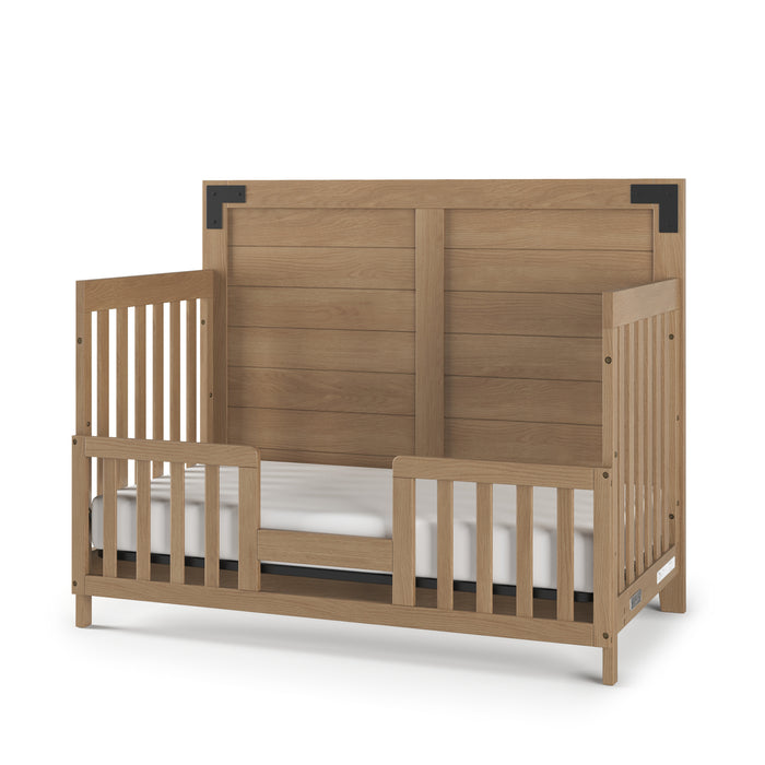 The Child Craft F09514 Toddler Guard Rail in Nutmeg features safety rails on both sides, an open front for easy access, and black metal accents on the headboard. Includes a white mattress for a safe and stylish crib transition.