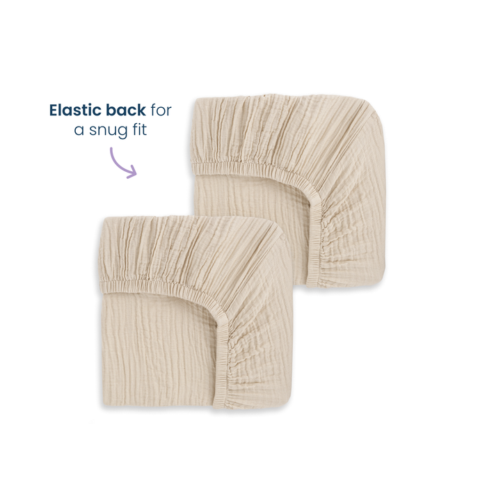 Two HALO Fitted Crib Sheets made from 100% organic cotton muslin are shown side by side. An arrow points to the elastic edge with text: "Elastic back for a snug fit." The 2-pack beige sheets sit on a clean white background.