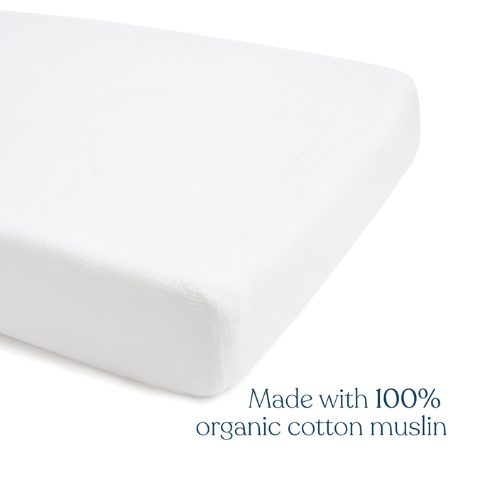 A close-up of a white, rectangular HALO Fitted Crib Sheet with text reading “Made with 100% organic cotton muslin”—perfect for gentle comfort and breathability; available in a 2-pack.