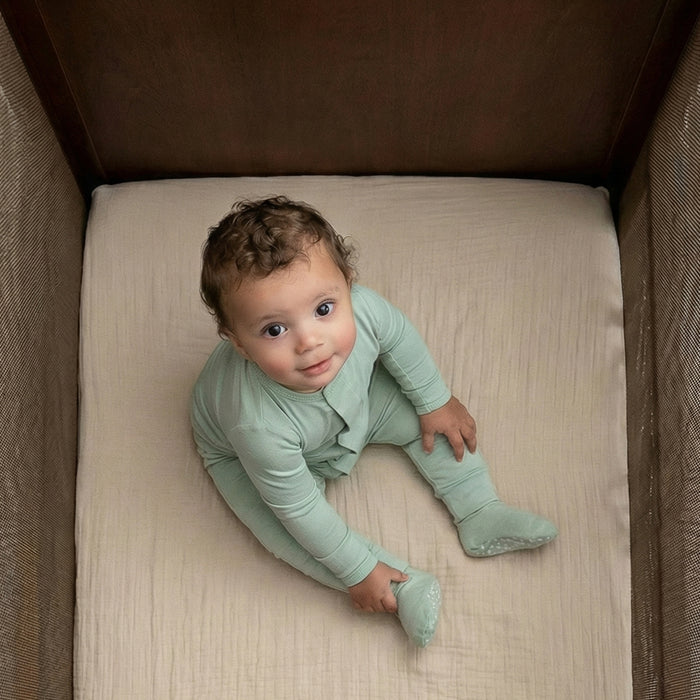 A baby in mint green pajamas sits in a crib with HALO Fitted Crib Sheets 100% Organic Cotton Muslin 2 Pack, gazing up at the camera with a curious expression.