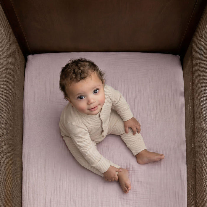 A baby with curly hair, in a light beige outfit, sits barefoot on HALO Fitted Crib Sheets 100% Organic Cotton Muslin 2 Pack in a wooden crib, gazing up at the camera with curiosity.