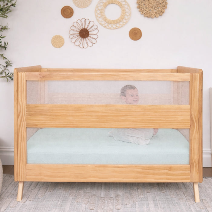 A happy baby sits in a wooden crib with mesh sides, outfitted with HALO Fitted Crib Sheets 100% Organic Cotton Muslin 2 Pack, in a softly lit room featuring woven wall hangings and a light rug.