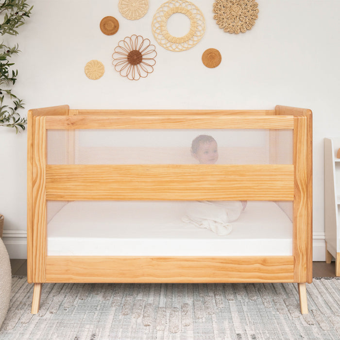 A baby sleeps in a wooden crib with mesh sides, wrapped in a white blanket. The crib is fitted with HALO Fitted Crib Sheets 100% Organic Cotton Muslin 2 Pack and is set in a bright room with woven wall decor and a patterned rug.