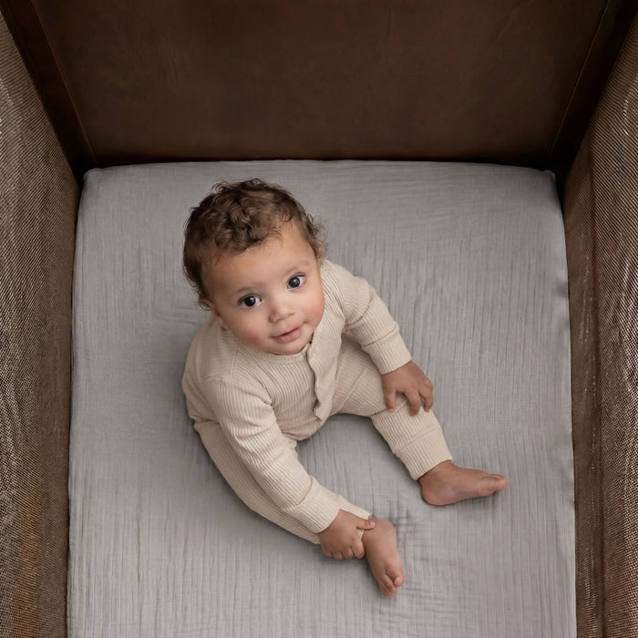 A baby with curly hair in a beige ribbed outfit sits barefoot on HALO Fitted Crib Sheets 100% Organic Cotton Muslin 2 Pack, inside a crib, gazing up at the camera with curiosity.