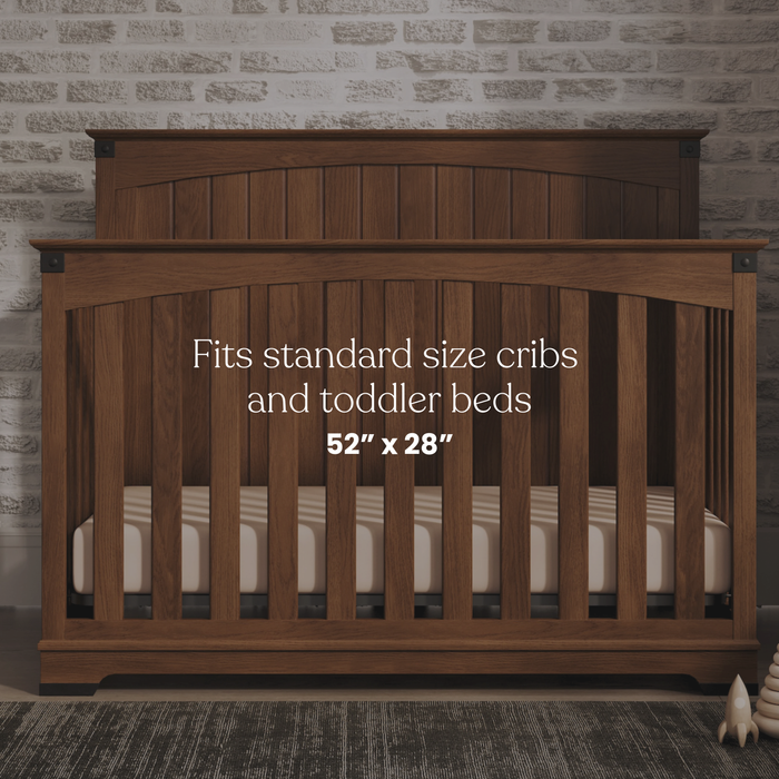 A wooden crib stands against a light brick wall. Featured: HALO Fitted Crib Sheets 100% Organic Cotton Muslin 2 Pack, fits standard 52 x 28 cribs and toddler beds. Add breathable, fitted sheets for comfort and style.
