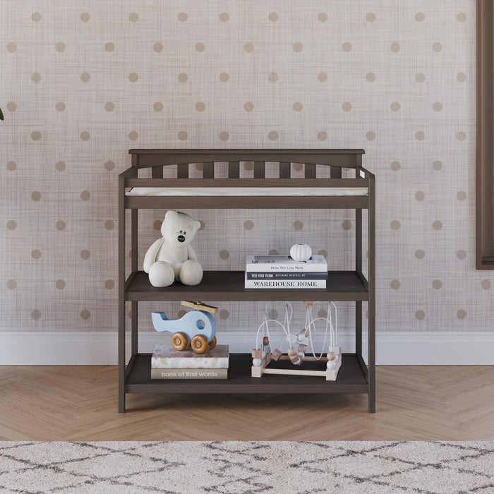The Child Craft Flat Top Baby Changing Table in dapper gray features a baby changing pad, shelves with a teddy bear, books, a blue elephant toy, and bead maze, set against a pink polka dot wall and patterned rug on wooden flooring.