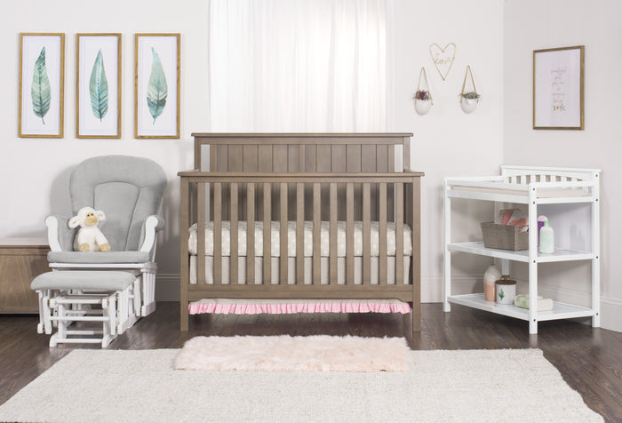 A cozy nursery with a wooden crib in baby-safe finishes, the Child Craft Flat Top Baby Changing Table in matte white for storage, a gray rocking chair with a stuffed lamb, leaf art, and soft decor accents on hardwood floors.