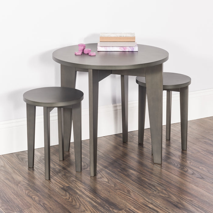 The Child Craft Geo Table and Stools Set in Dapper Gray features a small round table with two matching stools on a wooden floor. Stacked books and pink wooden blocks sit atop the table, set against a white wall with baseboard.