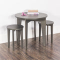 The Child Craft Geo Table and Stools Set in Dapper Gray features a small round table with two matching stools on a wooden floor. Stacked books and pink wooden blocks sit atop the table, set against a white wall with baseboard.