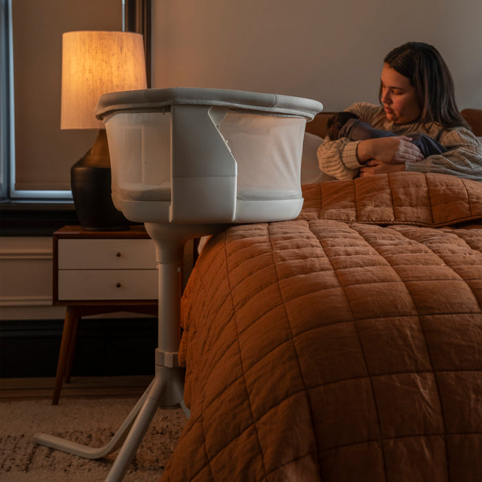 A woman sits on a bed with a rust quilt, holding a baby. Beside her is the white HALO® BassiNest® Swivel Sleeper 4.0 Signature by HALO. A lit lamp and nightstand are in the softly glowing background.