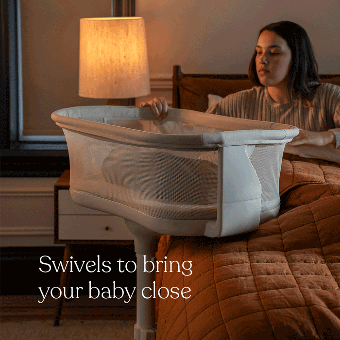 A woman sits on a bed beside the HALO® BassiNest® Swivel Sleeper 4.0 Signature, its mesh sides visible as she reaches for her baby while the 360° swivel brings the bassinet closer. Cozy lamp light glows. Text: Swivels to bring your baby close. Brand: HALO.