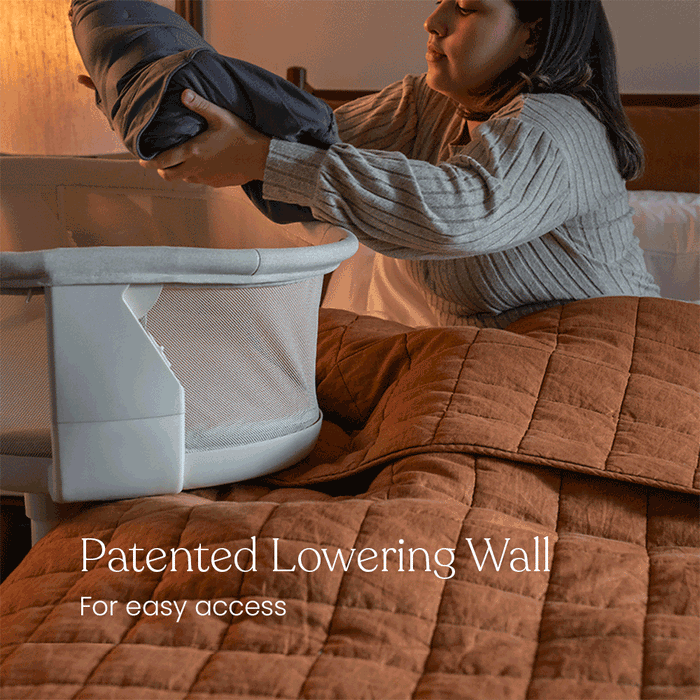 A woman in a gray sweater lowers the side wall of the HALO® BassiNest® Swivel Sleeper 4.0 Signature by HALO, holding a blanket, with “Patented Lowering Wall for easy access” over a brown quilted bedspread.