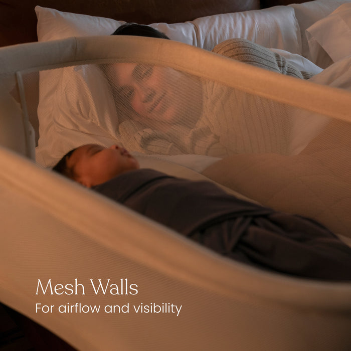 A parent relaxes in bed, watching their baby sleep in the HALO® BassiNest® Swivel Sleeper 4.0 Signature bassinet with mesh walls for airflow and visibility. Text on image: Mesh Walls – For airflow and visibility. Brand: HALO.