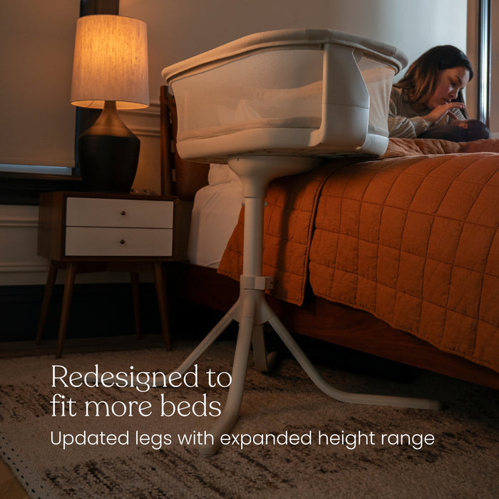 A HALO® BassiNest® Swivel Sleeper 4.0 Signature bassinet with updated legs stands by a bed with a rust quilt, while a woman sits on the bed using her phone. Text: Redesigned to fit more beds. Expanded height range for versatility.