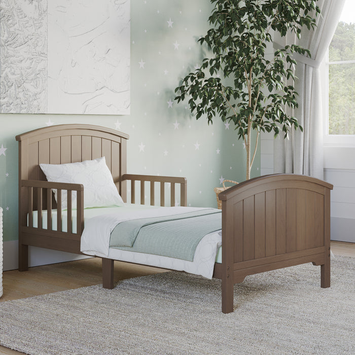 The Child Craft Hampton Toddler Bed in Dusty Heather is shown with a white pillow and bedding on a light gray rug in a softly decorated bedroom featuring star-patterned wallpaper, a potted tree, and curtained window.