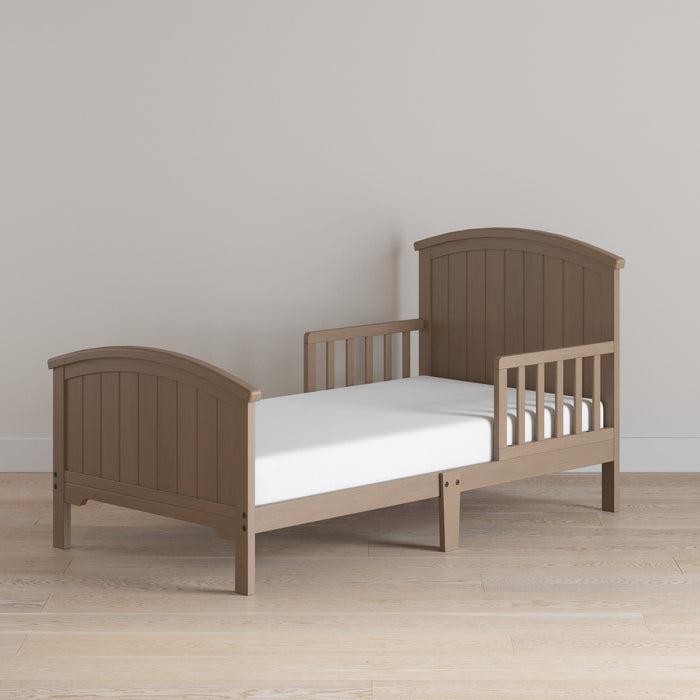 The Child Craft Hampton Toddler Bed in Dusty Heather, with side rails and a white mattress, sits on a light wood floor against a plain beige wall.