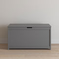 The Child Craft Harmony 33" Kids Toy Box in cool gray, featuring a flat lid and handle opening, is placed against a light-colored wall on a wooden floor.