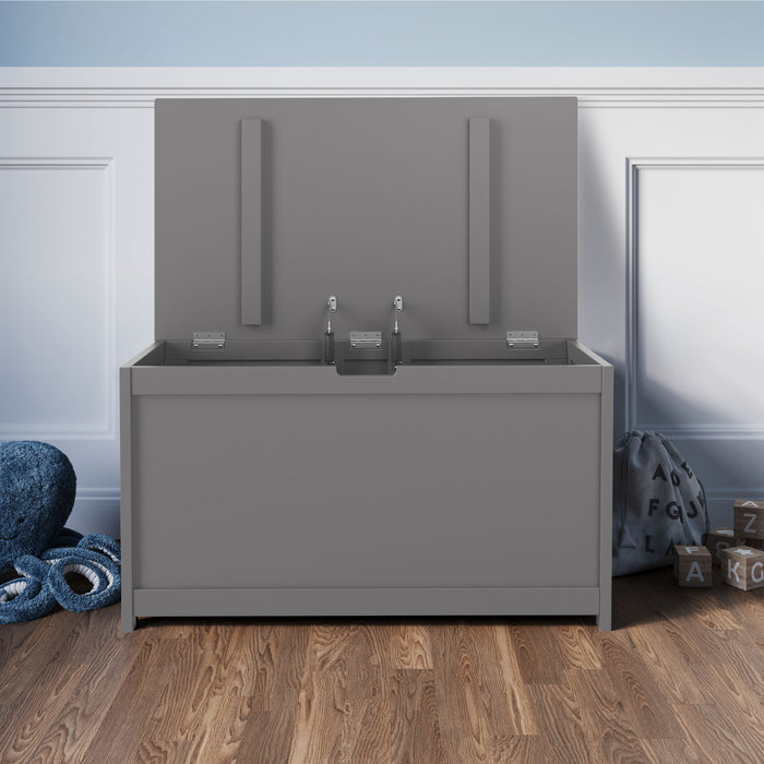 The Child Craft Harmony 33" Kids Toy Box in Cool Gray sits on a wooden floor by a blue wall, next to a plush blue rug, stuffed toy, fabric storage bag with letters, and wooden letter blocks.