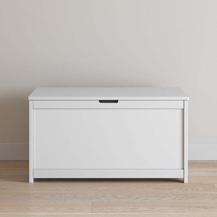 The Child Craft Harmony 33" Kids Toy Box in matte white with a hinged lid is placed against a beige wall on a light wood floor.