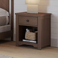 The Child Craft Harmony Nightstand in Dusty Heather, with child-safe finishes, has a drawer and open shelf for bedside storage. Below are a woven basket and CHANEL book; a square lamp sits on top by the bed and white shiplap wall.
