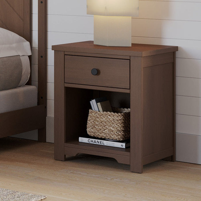 The Child Craft Harmony Nightstand in Dusty Heather, with child-safe finishes, has a drawer and open shelf for bedside storage. Below are a woven basket and CHANEL book; a square lamp sits on top by the bed and white shiplap wall.