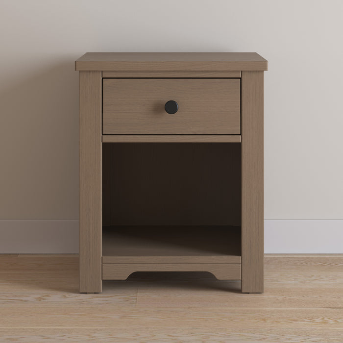 The Child Craft Harmony Nightstand in Dusty Heather features a single drawer with a round black knob, child-safe finishes, and an open shelf below—perfect for bedside storage. Pictured on a light wood floor against a plain beige wall.