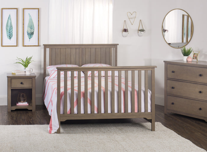 A cozy bedroom featuring a wooden bed frame, striped bedding, the Child Craft Harmony Nightstand in Dusty Heather with a plant on top, a dresser and round mirror, wall art, and soft white curtains that let in natural light.