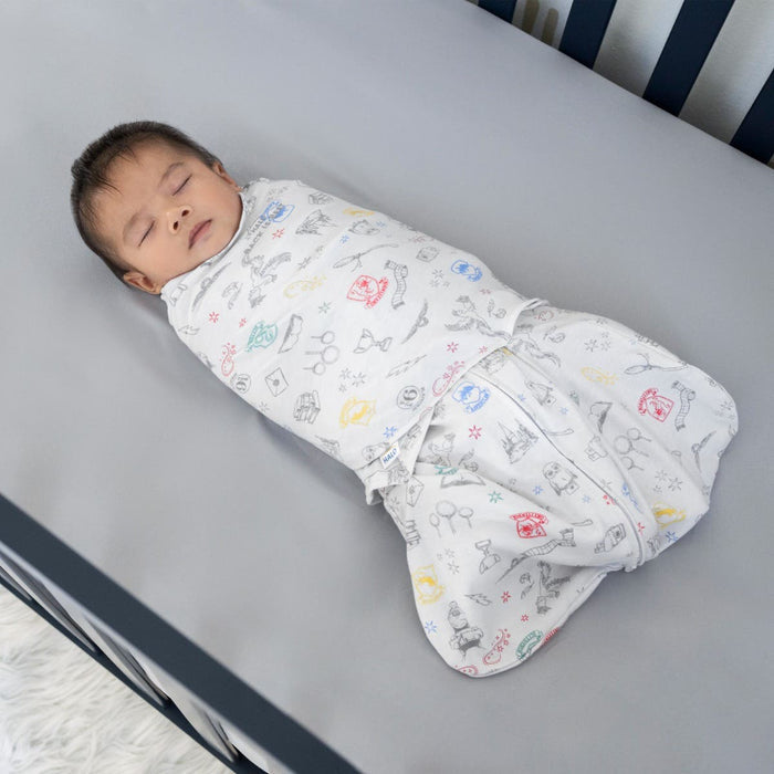 A baby sleeps peacefully in a crib, wrapped in the cozy HALO Harry Potter™ Swaddle made of 100% Cotton with a TOG rating of 1.5, adorned with colorful illustrations. The dark wooden frame contrasts beautifully with the light gray sheet, creating a magical sleep environment.