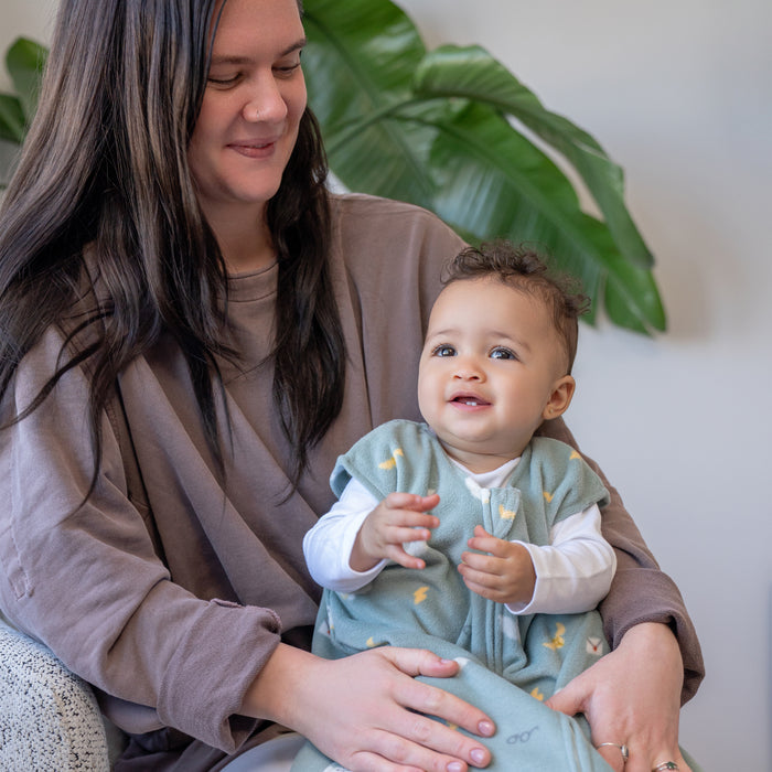 A woman with long dark hair smiles while holding a baby in a light blue patterned HALO Harry Potter™ Wearable Blanket Micro-Fleece TOG 1.0, sitting indoors with a large green leaf plant in the background.