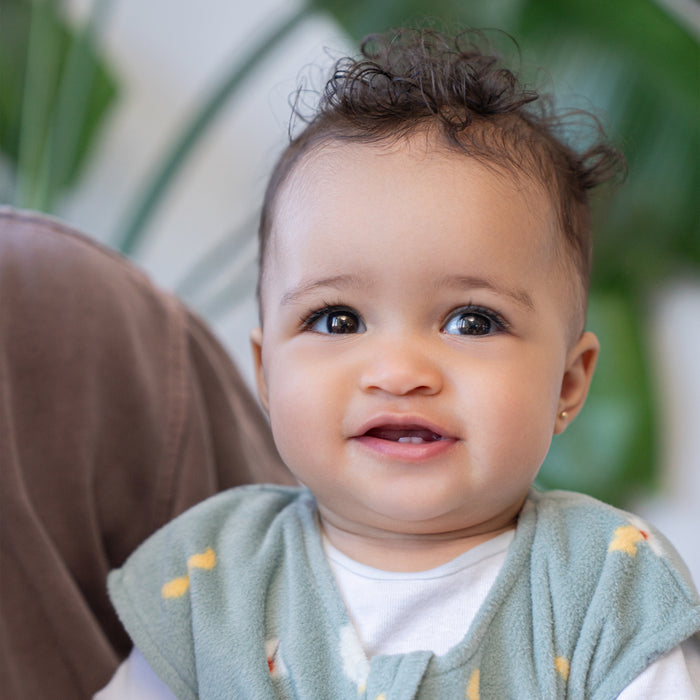 A smiling baby with curly hair and a light green bib with yellow dots sits next to an adult, cozy in the HALO Harry Potter™ Wearable Blanket Micro-Fleece TOG 1.0. Out-of-focus green plants add a fresh touch to the background.
