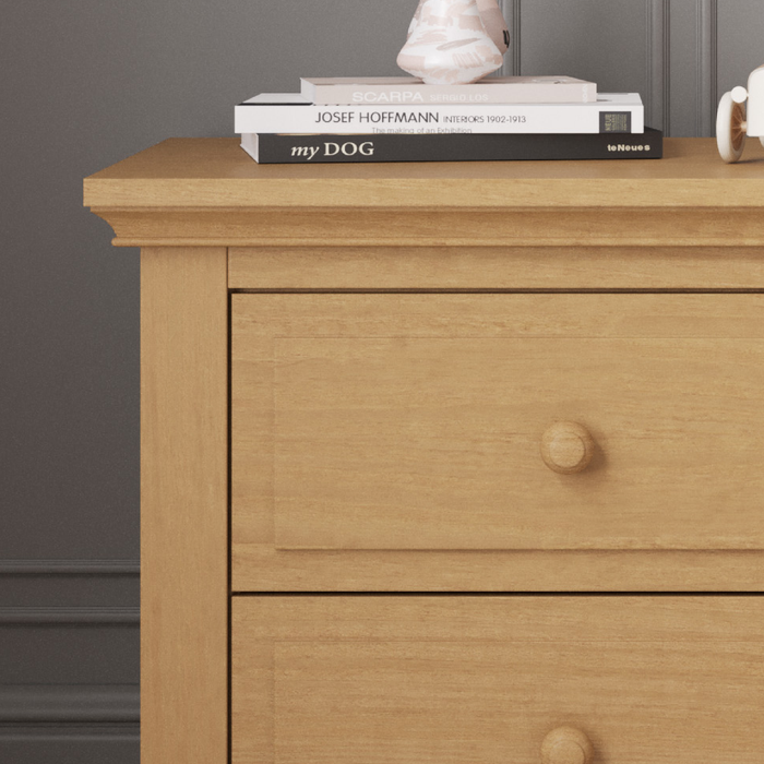 A Child Craft Jordyn Nightstand, Ready-to-Assemble in Biscotti, features three drawers. On top are two stacked books (one titled "my DOG"), a white mug, and a decorative object against a dark gray wall with crown molding.