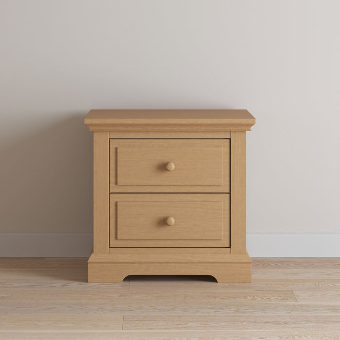 A Child Craft Jordyn Nightstand, Ready-to-Assemble in Biscotti, with two drawers and round knobs, stands on a light wood floor against a white wall—an ideal nursery nightstand.