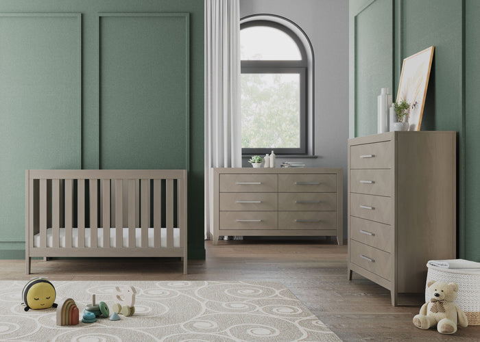 Modern nursery with green paneled walls, Child Craft Kieran 3-in-1 Convertible Crib in Crescent Gray, matching dresser and chest, round swirl-patterned rug, plush toys, rainbow toy, and a window with white curtains letting in natural light.