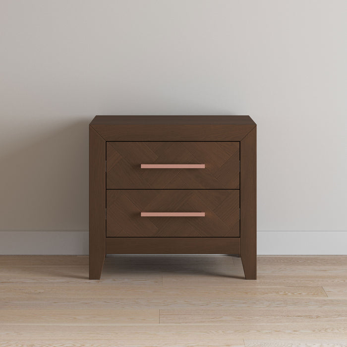 The Child Craft Kieran Nightstand in Toasted Chestnut, featuring two drawers with pink handles and baby-safe finishes, stands against a plain light-colored wall on a hardwood floor.