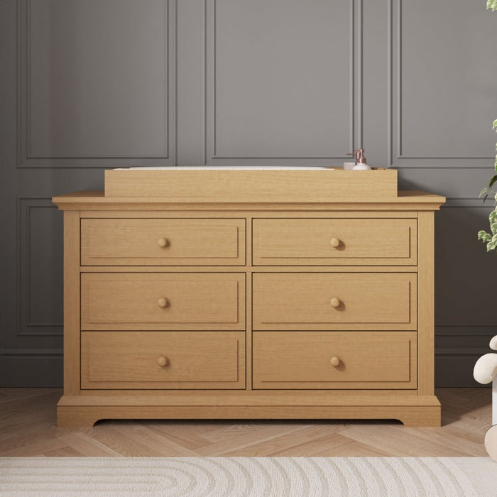 A Child Craft Legacy Changing Table Topper in Biscotti sits atop a six-drawer light wood dresser against a gray paneled wall. A small bottle, plus glimpses of a plant and soft toy, complete the cozy nursery scene.