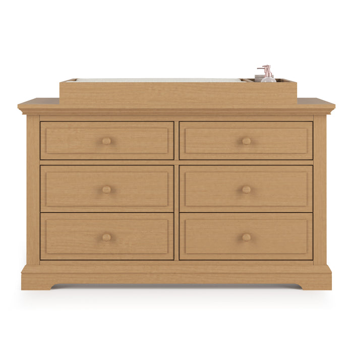 The Child Craft Legacy Changing Table Topper in Biscotti is placed on a wooden six-drawer dresser with round knobs, featuring a mat and a bottle of lotion or soap on the right side.