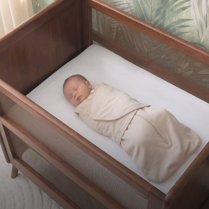 A newborn sleeps peacefully on their back in a HALO mini crib with Mini Crib Sheet 100% Cotton 2-Pack in white. They are swaddled in a beige blanket, with leafy wallpaper visible in the background.