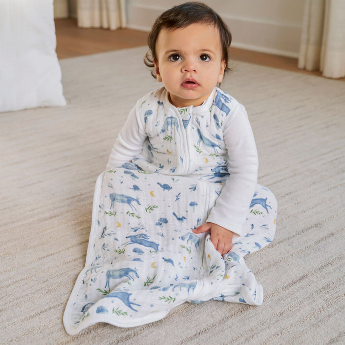 In a cozy room, a toddler sits on a light-colored carpet, snug in an aden + anais organic cotton light sleep sack TOG 1.0 with a zipper. The white and blue patterned sack boasts delightful animal illustrations, while the inquisitive child gazes upward.