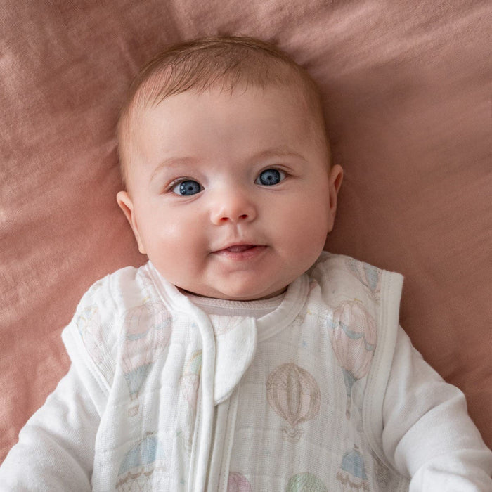 A baby with bright blue eyes and light brown hair lies content on a soft pink blanket, wrapped in an aden + anais organic cotton light sleep sack TOG 1.0. The white outfit, decorated with pastel hot air balloons, complements the GOTS certified 100% organic cotton muslin, showcasing the babys slight smile.