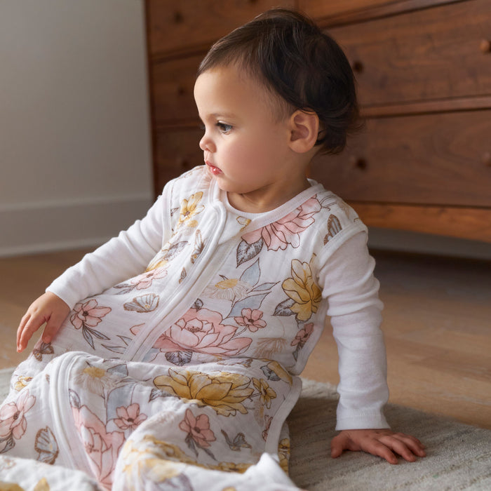 A toddler sits calmly on a carpeted floor next to a wooden dresser, wearing an aden + anais organic cotton light sleep sack TOG 1.0, adorned with a floral pattern. The soft lighting enhances the rooms warmth and highlights the comfort of the GOTS certified 100% organic cotton muslin.