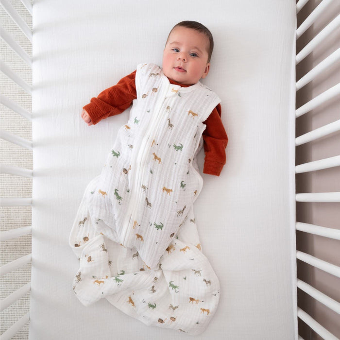 A baby wearing an aden + anais organic cotton light sleep sack TOG 1.0 with colorful animal prints lies in a crib. The baby, in a red long-sleeve shirt, looks up calmly. The crib features white bars and a cozy 100% organic cotton muslin mattress.