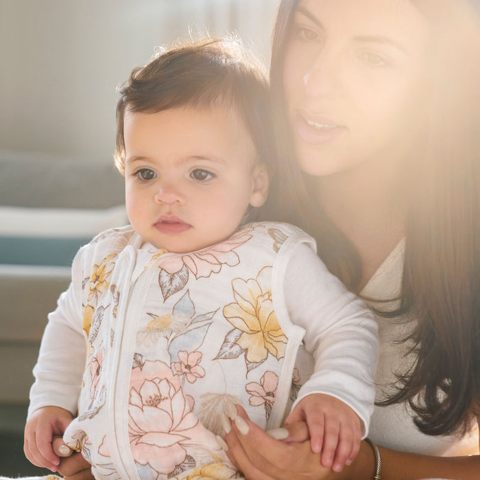 A woman cradles a baby in her arms as they gaze forward. The baby, dressed in a floral outfit, is cozy in an aden + anais organic cotton light sleep sack TOG 1.0. Sunlight streams through, casting a warm, soft glow behind them.