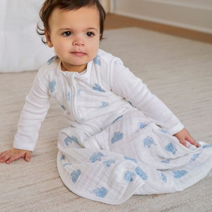 A toddler in a white outfit with blue patterns sits on a light carpet, snug in an aden + anais organic cotton light sleep sack TOG 1.0. With short, dark hair, the child looks slightly to the side in a neutrally toned room with minimal decor.