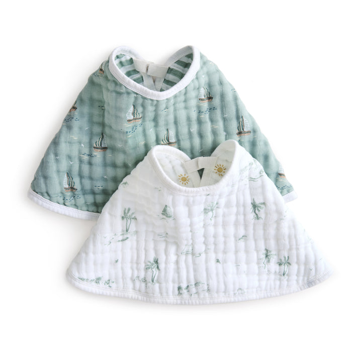 Displayed are two aden + anais organic cotton muslin burpy bibs: a green one with a nautical sailboat design and a white one featuring palm tree and island illustrations. Both have rounded necklines and fasten at the back.