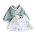 Displayed are two aden + anais organic cotton muslin burpy bibs: a green one with a nautical sailboat design and a white one featuring palm tree and island illustrations. Both have rounded necklines and fasten at the back.