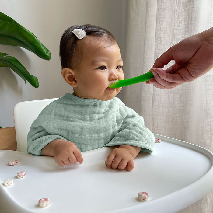 A baby in a high chair, wearing a light green bib, is fed with a green spoon. Snacks lie scattered on the tray in this bright and minimal setting, highlighting aden + anais organic cotton muslin burpy bibs 2 pack with plants and curtains as background décor.