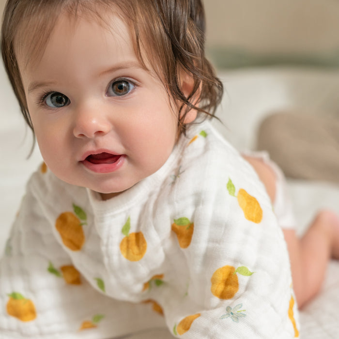 A baby with brown hair and blue eyes crawls on a soft surface, wearing a white outfit with yellow and orange fruit patterns and an aden + anais organic cotton muslin burpy bib from the 2 pack, looking curiously at the camera.