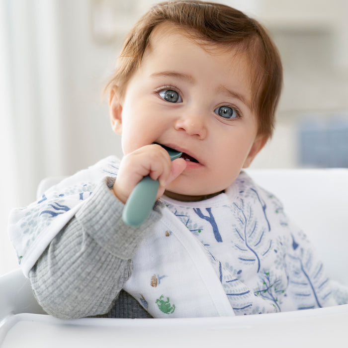 A baby with brown hair and blue eyes sits in a high chair, wearing a patterned bib from the aden + anais Organic Baby Essentials Bundle. Holding a spoon to their mouth, the baby looks curiously at the camera amidst a softly blurred background.