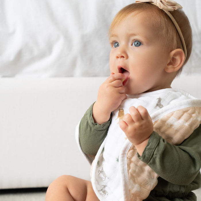 A baby with light brown hair and a pink bow sits curiously, wearing a green outfit and patterned bib from the HALO Welcome Home Bundle, with one hand near their mouth. Theyre surrounded by newborn essentials on a cozy white blanket or cushion.