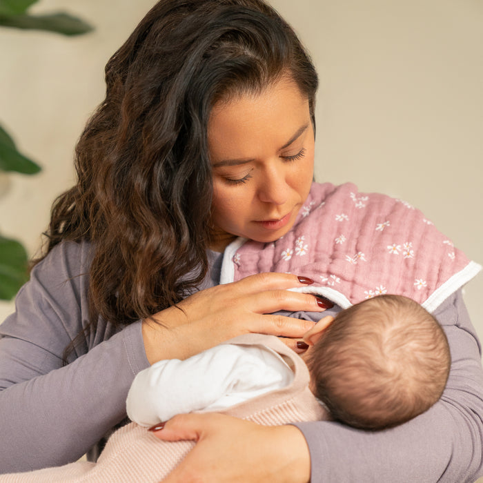 A woman with long dark hair cradles a baby, holding an aden + anais organic cotton muslin burpy bibs 2 pack over her shoulder. The soft background puts the focus on the cozy bib and tender moment.